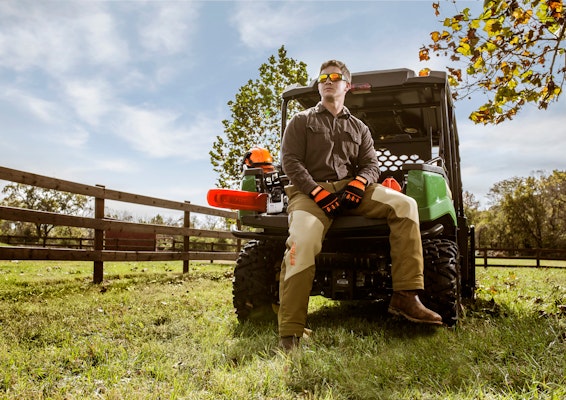 Man wearing Performance Pants from STIHL sitting on the back of a John Deere Utility Vehicle next to a chainsaw.