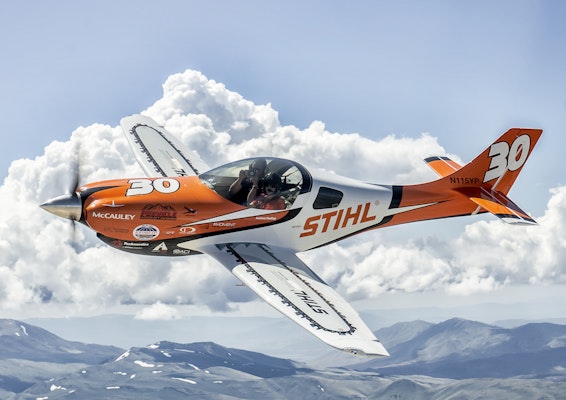 A STIHL Air Racing plane soaring past clouds in a blue sky over mountains 