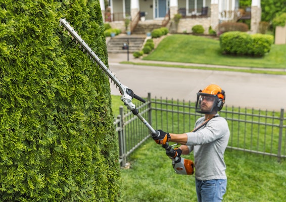 Man using the HLA 135 K (145°) to trim some hedges with a fence and house in the background.